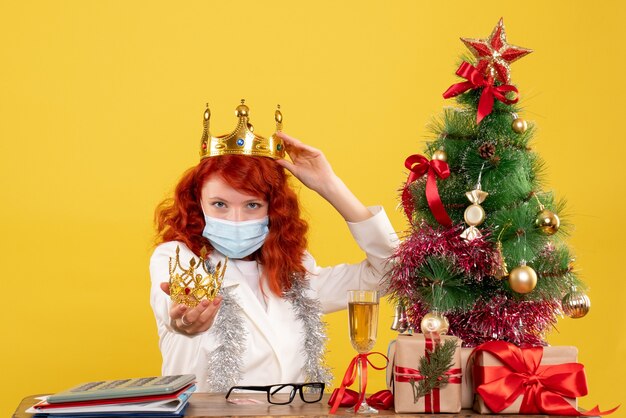 Front view female doctor sitting with xmas presents and holding crown on yellow desk