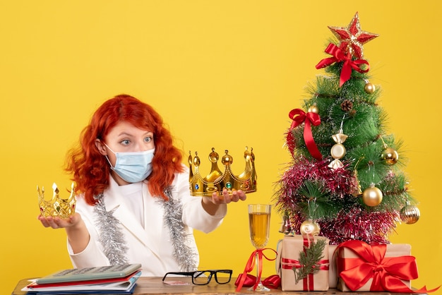 Front view female doctor sitting with xmas presents and holding crowns on yellow background