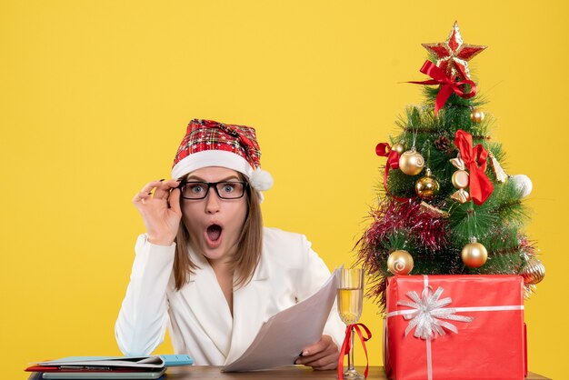 Front view female doctor sitting with xmas presents holding documents on a yellow background