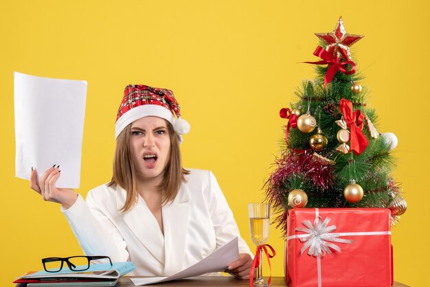 Вид спереди женщина-врач, сидящая с рождественскими подарками, держит документы на желтом фоне