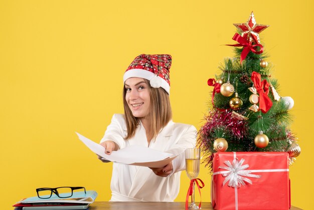 Вид спереди женщина-врач, сидящая с рождественскими подарками, держит документы на желтом фоне