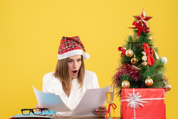 Front view female doctor sitting with xmas presents holding documents on yellow desk