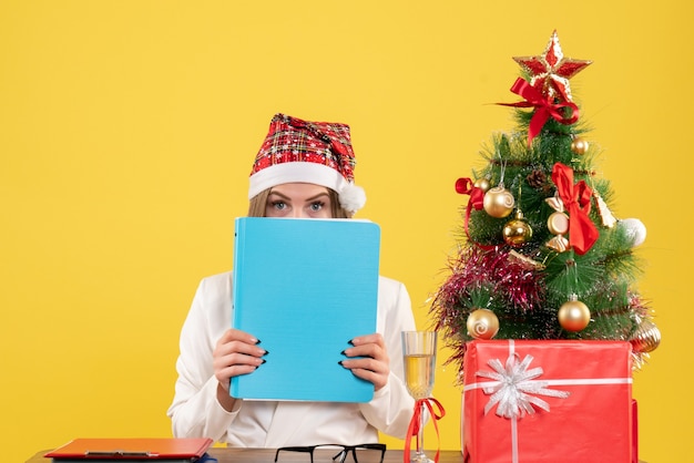 Вид спереди женщина-врач, сидящая с рождественскими подарками, держит файлы на желтом фоне