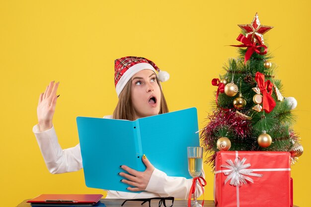 Front view female doctor sitting with xmas presents holding files on the yellow background