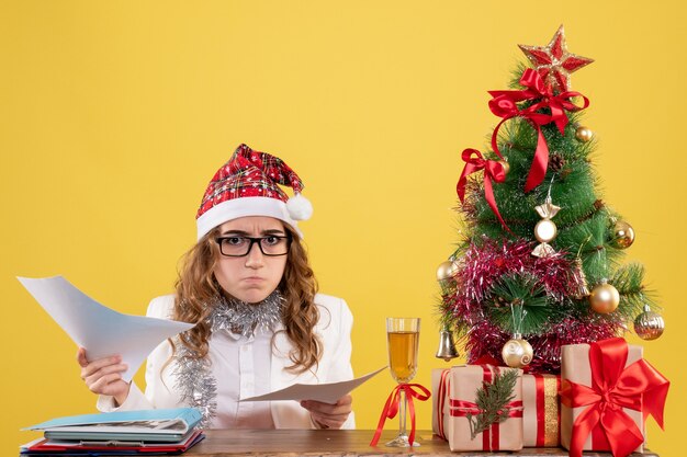 Free photo front view female doctor sitting with xmas presents tree and holding documents on a yellow background