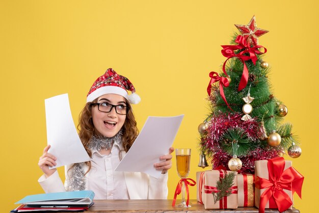 Front view female doctor sitting with xmas presents tree and holding documents on a yellow background
