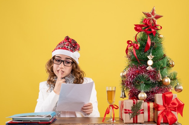 Front view female doctor sitting with xmas presents tree and holding documents on a yellow background