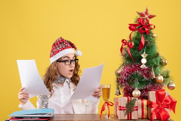 Free photo front view female doctor sitting with xmas presents tree and holding documents on yellow background