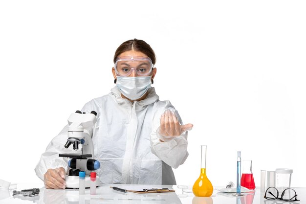 Front view female doctor in special suit and wearing mask using microscope and calling someone on white desk covid- virus pandemic coronavirus