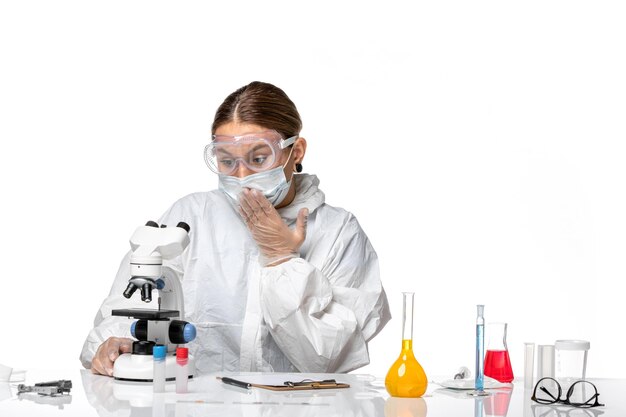 Front view female doctor in special suit and wearing mask using microscope and working on white background covid- virus pandemic coronavirus
