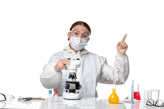 Front view female doctor in special suit and wearing mask working with microscope on a light white background covid virus pandemic coronavirus