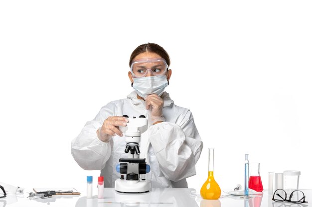 Вид спереди женщина-врач в специальном костюме и в маске, работающая с микроскопом и думающая на белом фоне, пандемия коронавируса вируса ковид