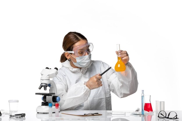 특수 양복과 흰색 책상에 노란색 솔루션으로 플라스크를 들고 마스크가있는 전면보기 여성 의사 covid pandemic coronavirus virus
