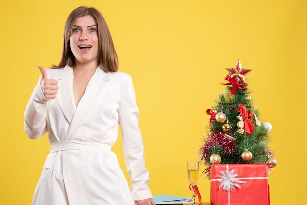 Front view female doctor standing around table with little christmas tree on a yellow background with christmas tree and gift boxes