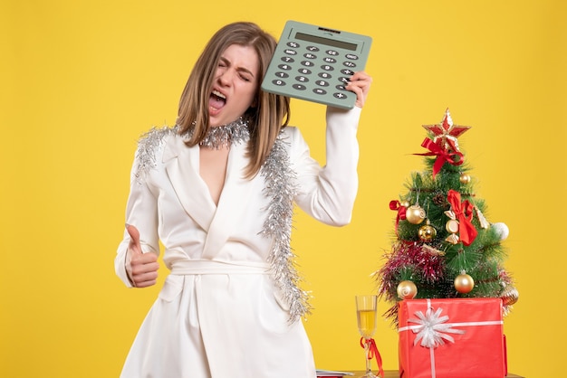 Front view female doctor standing and holding calculator on yellow desk with christmas tree and gift boxes