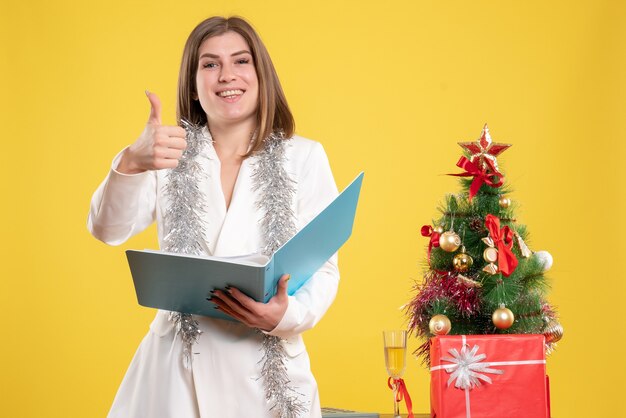 Front view female doctor standing and holding documents on yellow desk with christmas tree and gift boxes