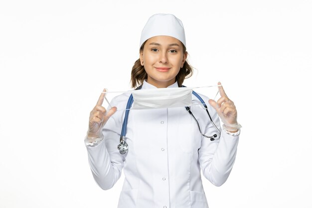 Front view female doctor in white medical suit holding mask on white wall pandemic disease medicine virus isolation