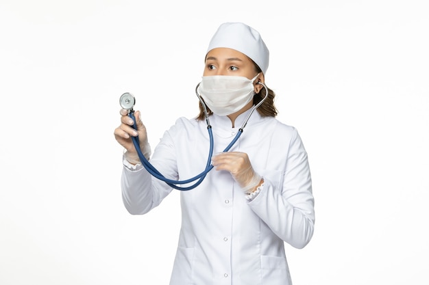 Front view female doctor in white medical suit and mask checking heart condition with stethoscope on white desk pandemy virus disease illness medicine