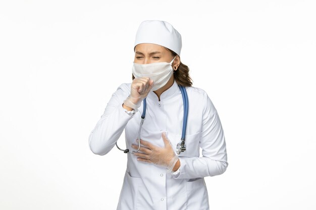 Front view female doctor in white medical suit and mask coughing up on white wall pandemy virus disease illness medicine