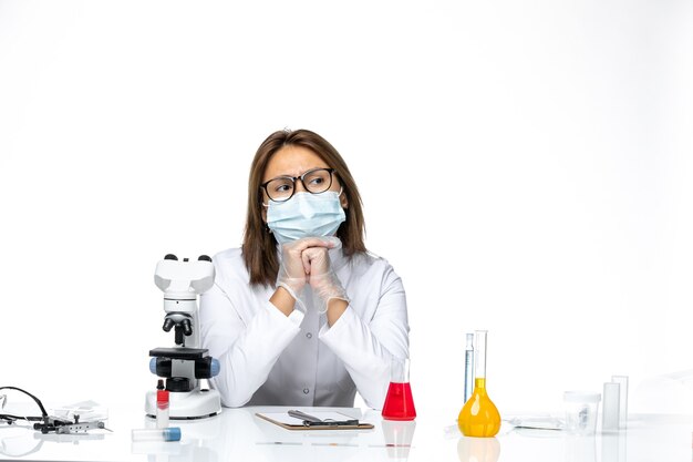 Front view female doctor in white medical suit and mask due to coronavirus sitting with solutions on white space