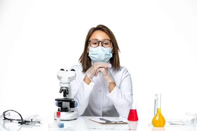 Front view female doctor in white medical suit and mask due to coronavirus sitting with solutions on white space