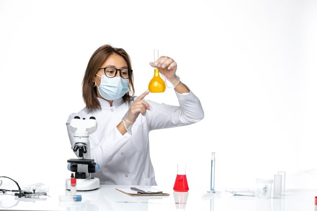 Front view female doctor in white medical suit and mask due to coronavirus working with solutions on a white space