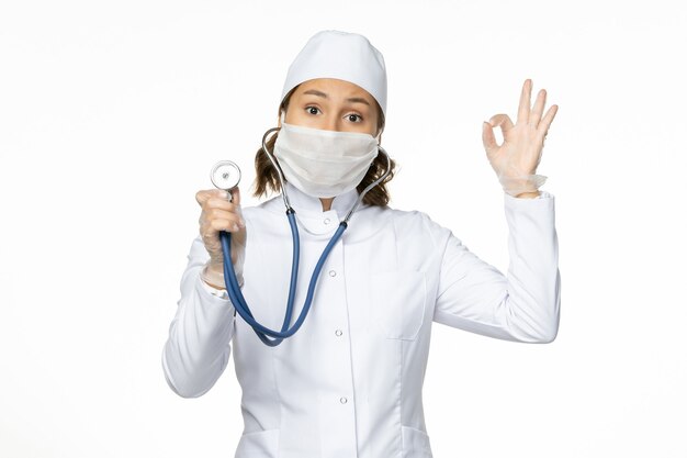 Вид спереди женщина-врач в белом медицинском костюме и маске, использующая стетоскоп на светлой белой стене, медицина болезни пандемического вируса