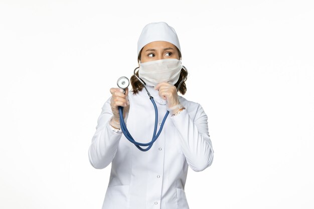 Front view female doctor in white medical suit and mask using stethoscope and thinking on white wall pandemy virus disease medicine