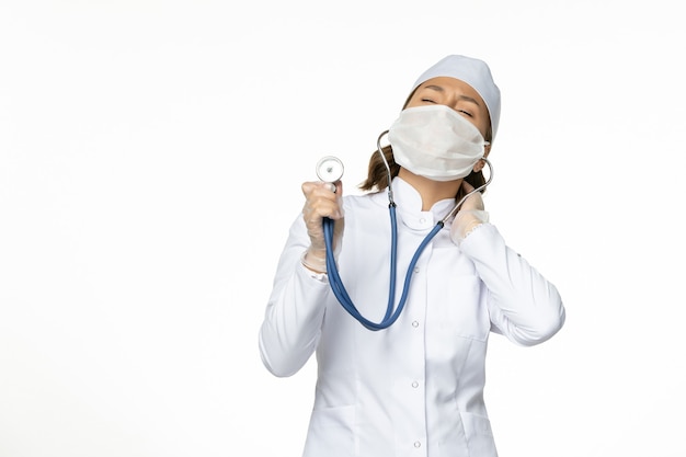 Front view female doctor in white medical suit and mask using stethoscope on the white desk pandemic disease medicine virus isolation