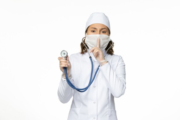 Front view female doctor in white medical suit and mask using stethoscope on the white desk pandemic virus disease medicine