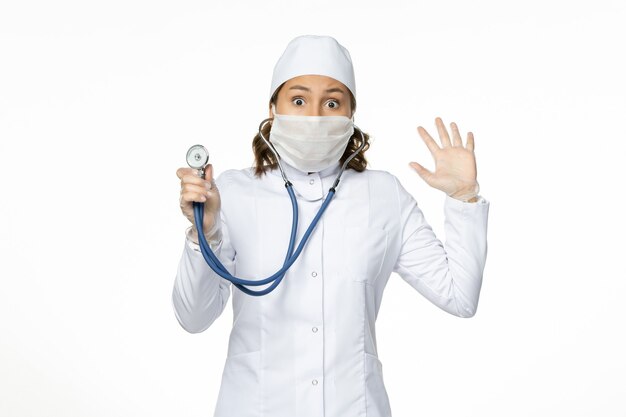 Front view female doctor in white medical suit and mask using stethoscope on white wall pandemic viruses disease medicine