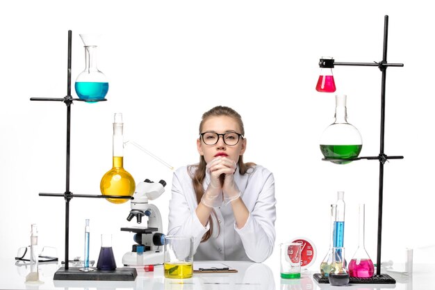 Front view female doctor in white medical suit sitting in front of table with solutions on a white background covid chemistry virus pandemic