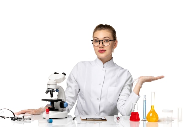 Front view female doctor in white medical suit sitting in front of table with solutions on a white desk