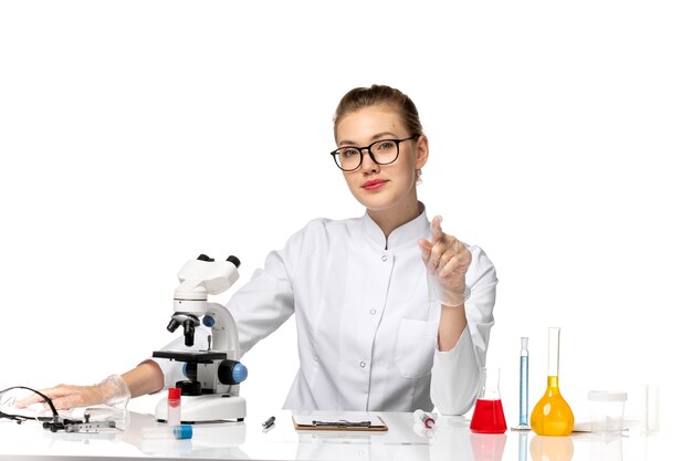 Front view female doctor in white medical suit sitting in front of table with solutions on the white space