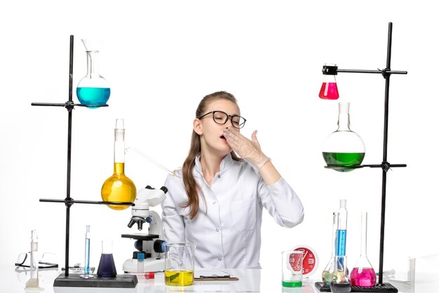 Вид спереди женщина-врач в белом медицинском костюме, сидящая перед столом с решениями, зевая на белом полу, пандемия вируса химии ковид