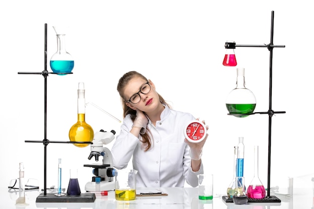 Front view female doctor in white medical suit sitting and holding clocks on a white background virus chemistry pandemic covid