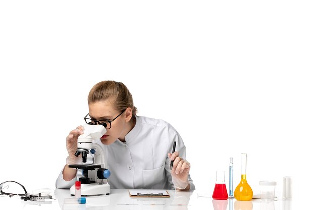 Front view female doctor in white medical suit using microscope on a white space