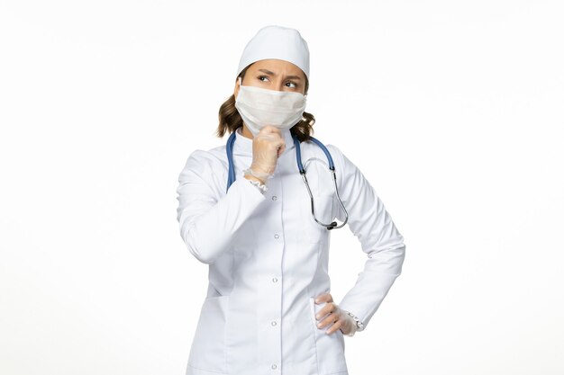 Front view female doctor in white medical suit and wearing mask due to coronavirus thinking on white wall pandemic disease isolation covid