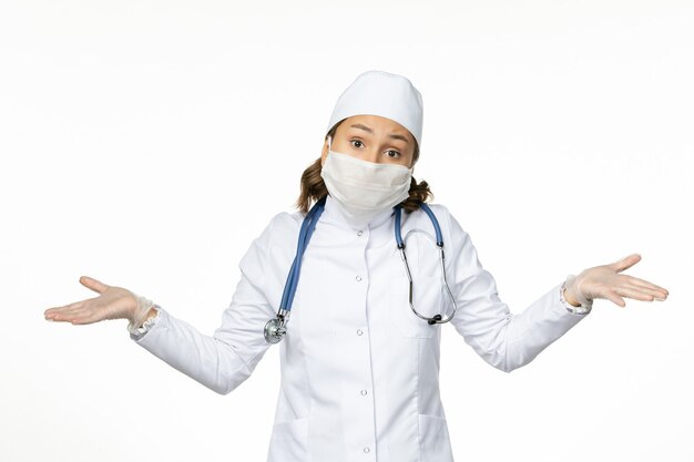 Front view female doctor in white medical suit and wearing mask due to coronavirus on the white wall pandemic virus disease isolation covid