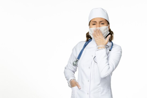 Вид спереди женщина-врач в белом медицинском костюме и в маске из-за коронавируса, зевая на белой стене, изоляция пандемического заболевания covid