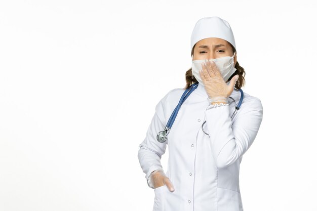 Front view female doctor in white medical suit and wearing mask due to coronavirus yawning on white wall pandemic disease isolation covid