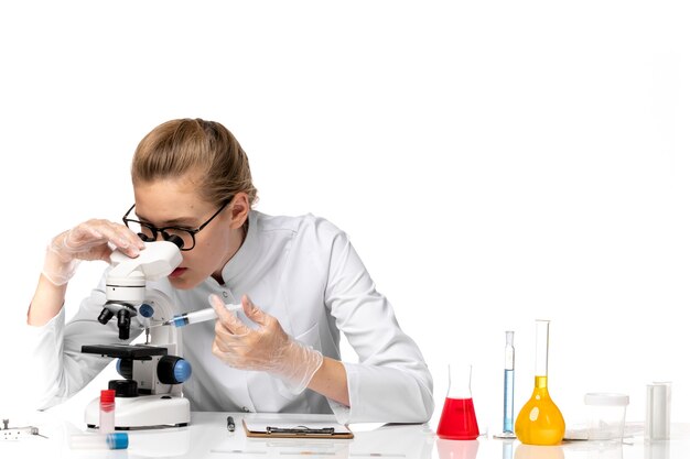 Front view female doctor in white medical suit with gloves holding injection and using microscope on a white space