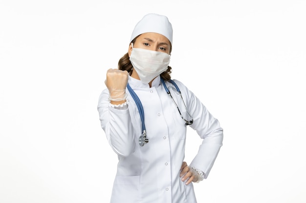 밝은 흰색 책상에 코로나 바이러스로 인해 흰색 의료 복과 마스크가있는 전면보기 여성 의사 대유행 바이러스 질병 격리 COVID