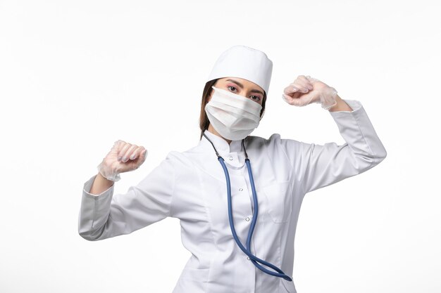 コロナウイルスが白い壁の病気の薬のウイルスのパンデミックコビッドでわずかに踊っているため、マスクをした白い医療スーツを着た正面図の女性医師-