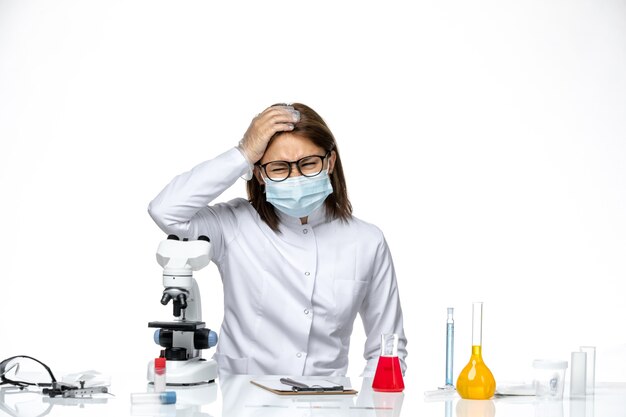 Front view female doctor in white medical suit with mask due to covid sitting on light white space