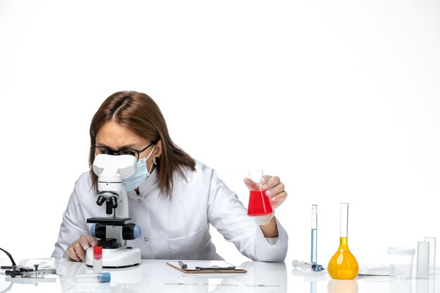 Front view female doctor in white medical suit with mask due to covid using microscope on light-white space