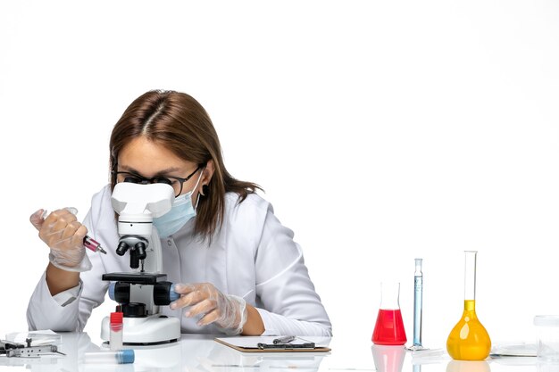 Front view female doctor in white medical suit with mask due to covid using microscope on a white space