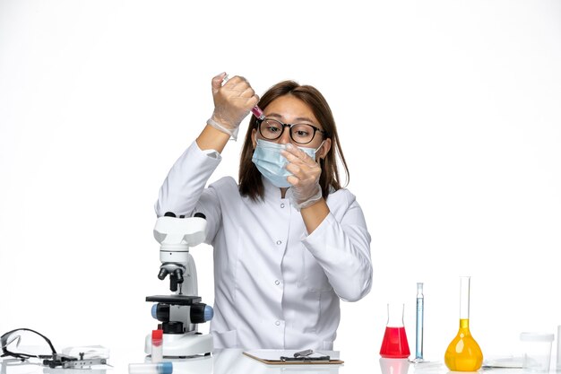 Front view female doctor in white medical suit with mask due to covid working with solutions on the white space