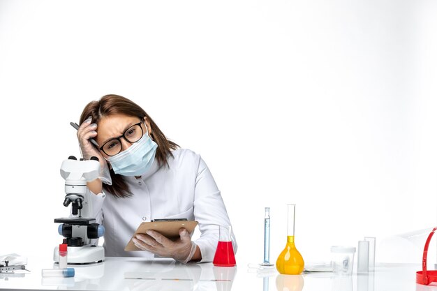 Front view female doctor in white medical suit with mask due to covid writing something on light white space