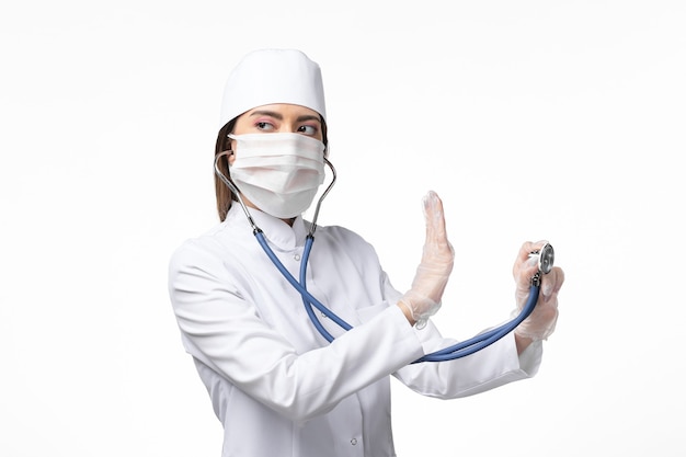 Front view female doctor in white medical suit with a mask due to pandemic holding a stethoscope on white desk disease medicine virus pandemic covid-